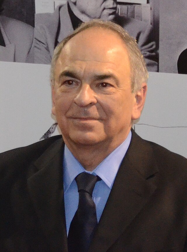 Gabriel Liiceanu in a suit and tie, standing in front of a grayscale photograph, captured by Razvan Socol.