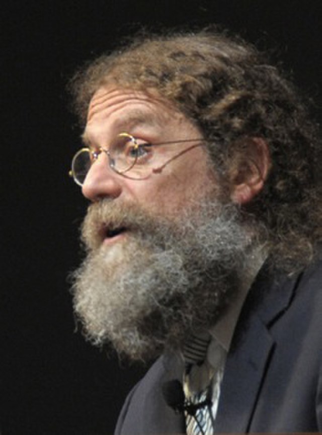 Robert Sapolsky speaking on stage, wearing a suit and tie with a lapel microphone, during a public lecture.