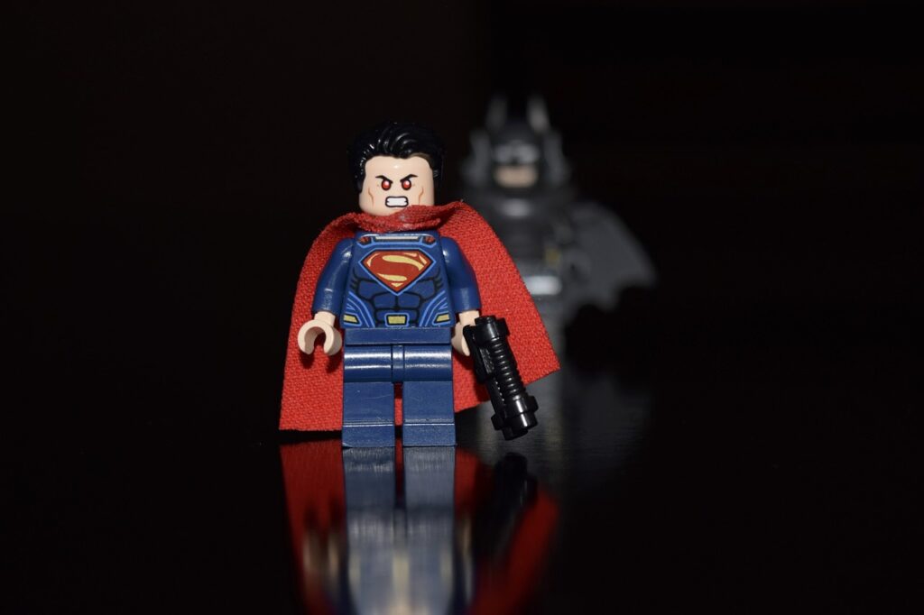 "LEGO Superman minifigure in the foreground with a red cape and blue suit, standing on a reflective surface, with a LEGO Batman minifigure blurred in the background under dramatic low-key lighting